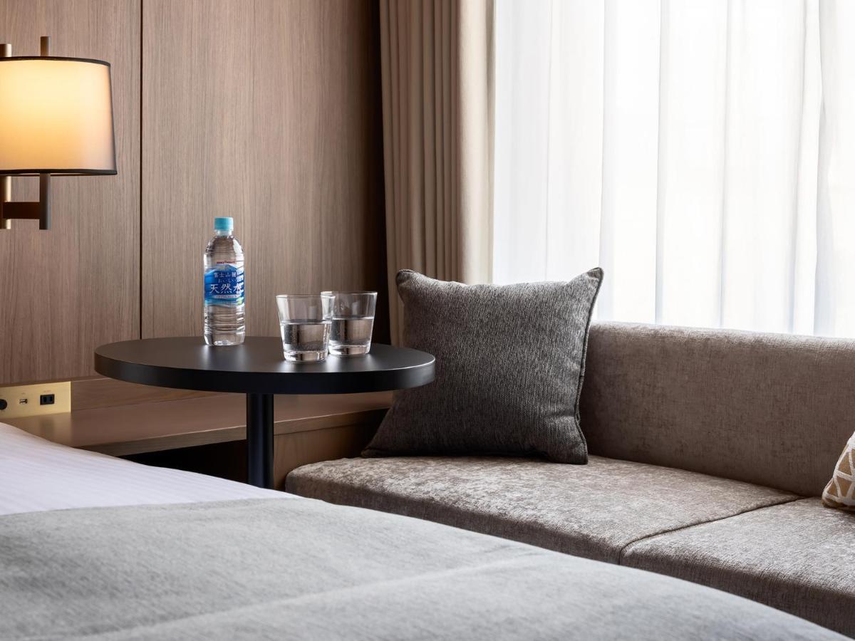 Mitsui Garden Hotel Roppongi Tokyo Premier Esterno foto The photo depicts a cozy hotel room setting. On the left, there is a bedside table with a lamp. Against the wall, there is a comfortable-looking couch with a decorative pillow. In front of the couch, there's a small round table that holds a bottle of