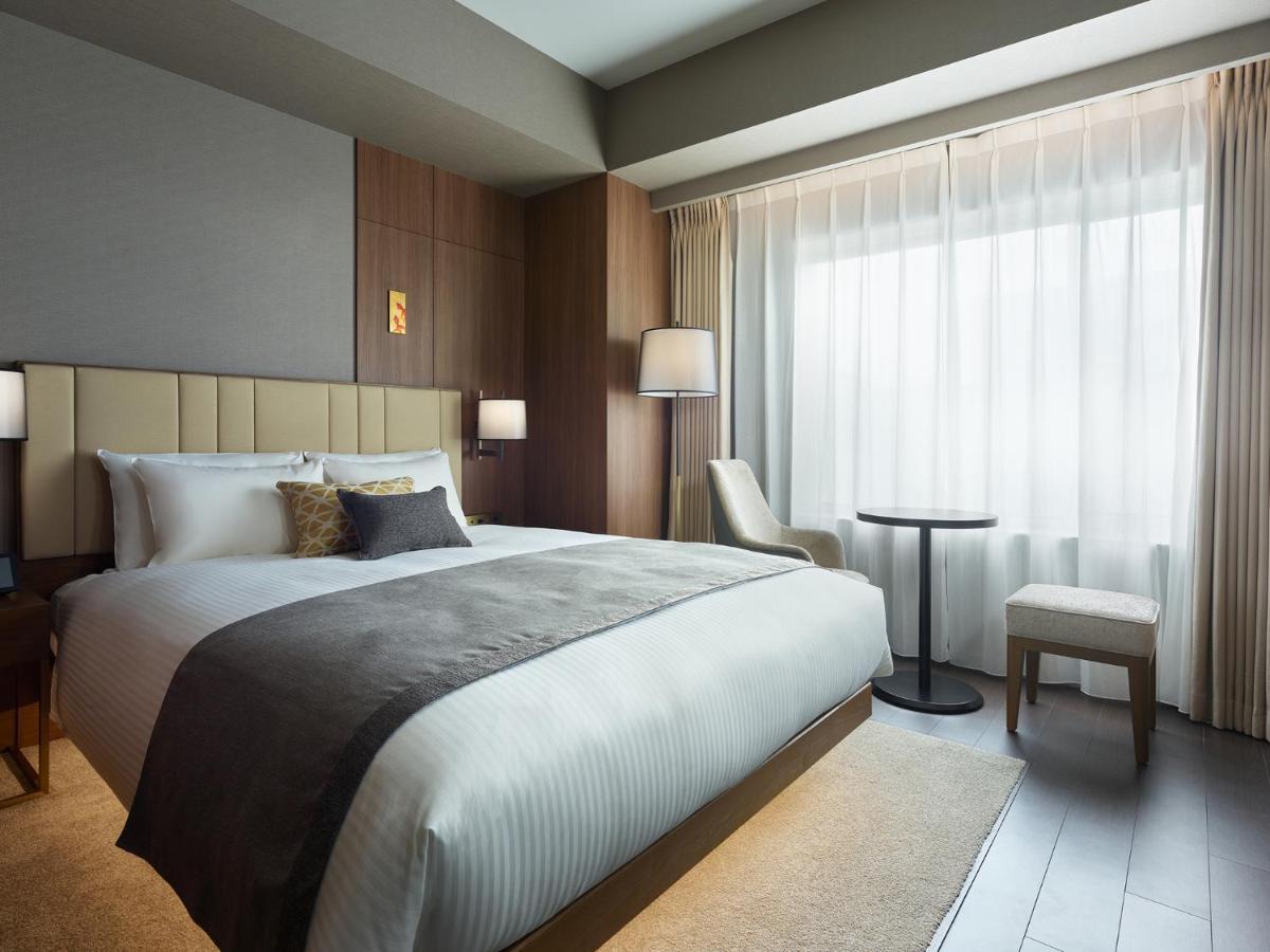Mitsui Garden Hotel Roppongi Tokyo Premier Esterno foto The photo shows a modern hotel room featuring a comfortable bed with white linens and a decorative throw pillow. The room has a neutral color palette with wooden accents on the walls. There are two bedside lamps and a chair positioned next to a small