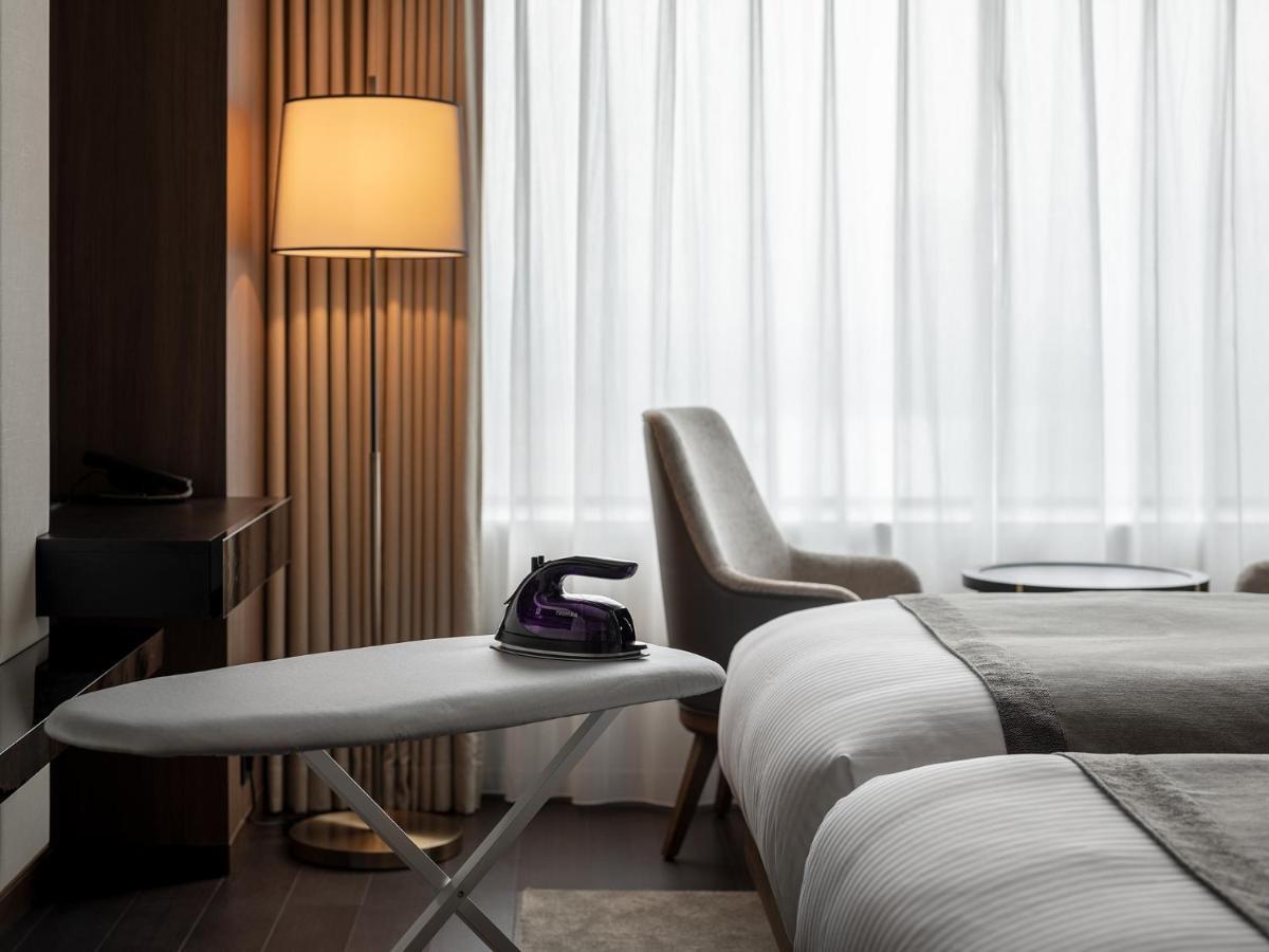 Mitsui Garden Hotel Roppongi Tokyo Premier Esterno foto The photo shows a modern hotel room featuring two beds with white bedding. There is a light-colored armchair next to a small side table. On the table, there is an iron, and beside it, a lamp with a warm glow is illuminating the space. The large windo