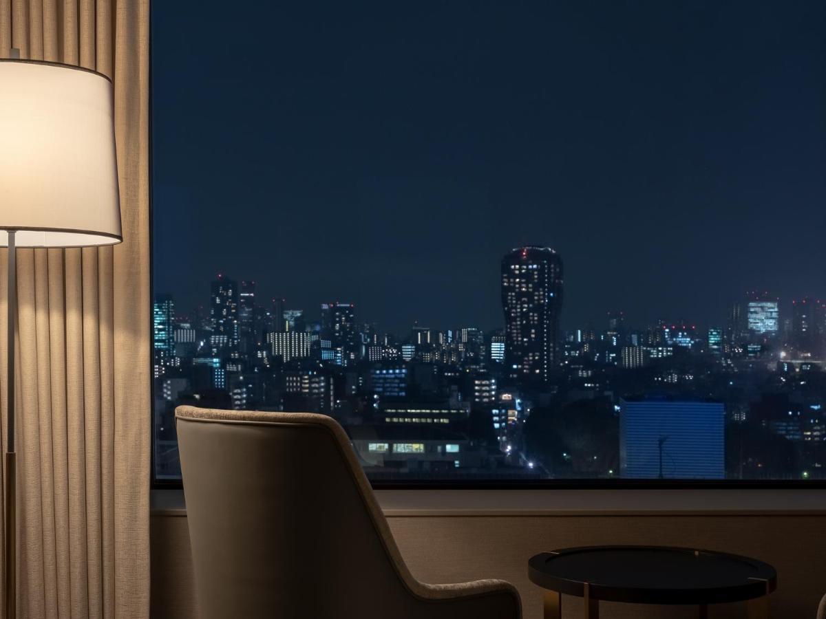 Mitsui Garden Hotel Roppongi Tokyo Premier Esterno foto The photo depicts a cozy hotel room setting at night. In the foreground, there's a stylish armchair next to a small round table. A standing lamp provides soft lighting, creating a warm ambiance. In the background, large windows reveal a city skyline 