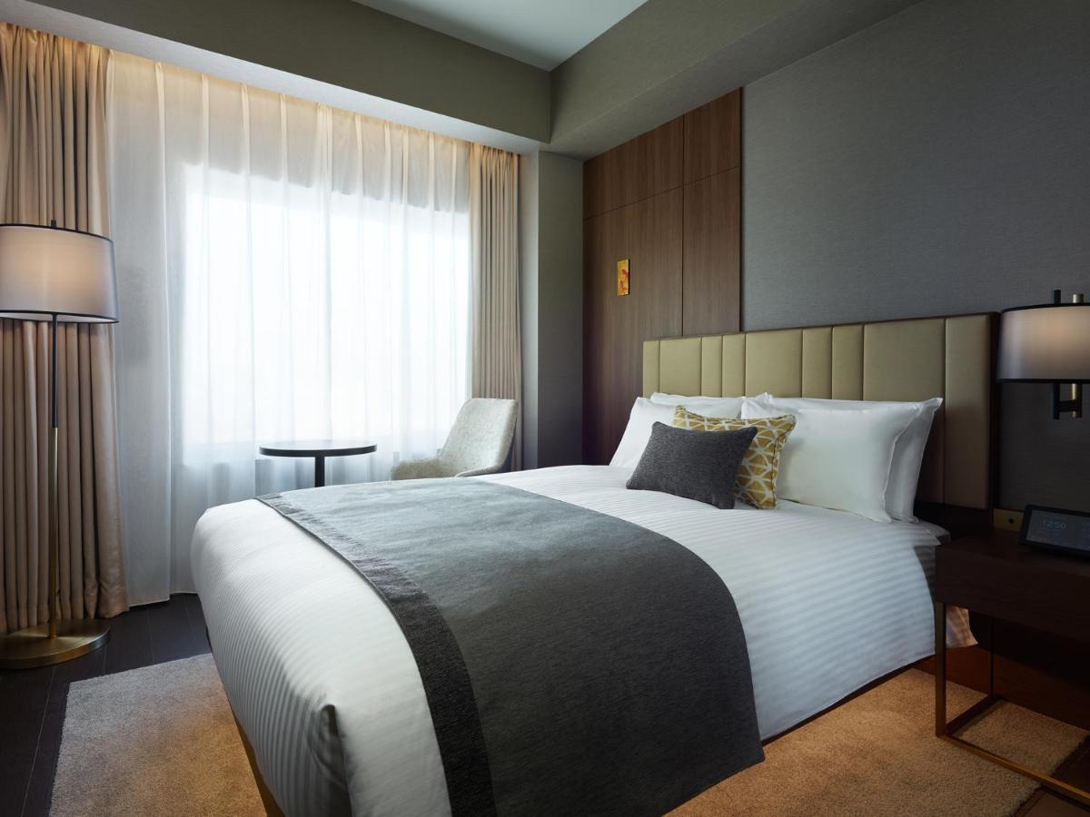 Mitsui Garden Hotel Roppongi Tokyo Premier Esterno foto The photo shows a modern hotel room featuring a neatly made bed with white linens and a decorative gray throw blanket. To the side, there is a small round table and a comfortable chair, creating a cozy seating area. The room is well-lit with natural 