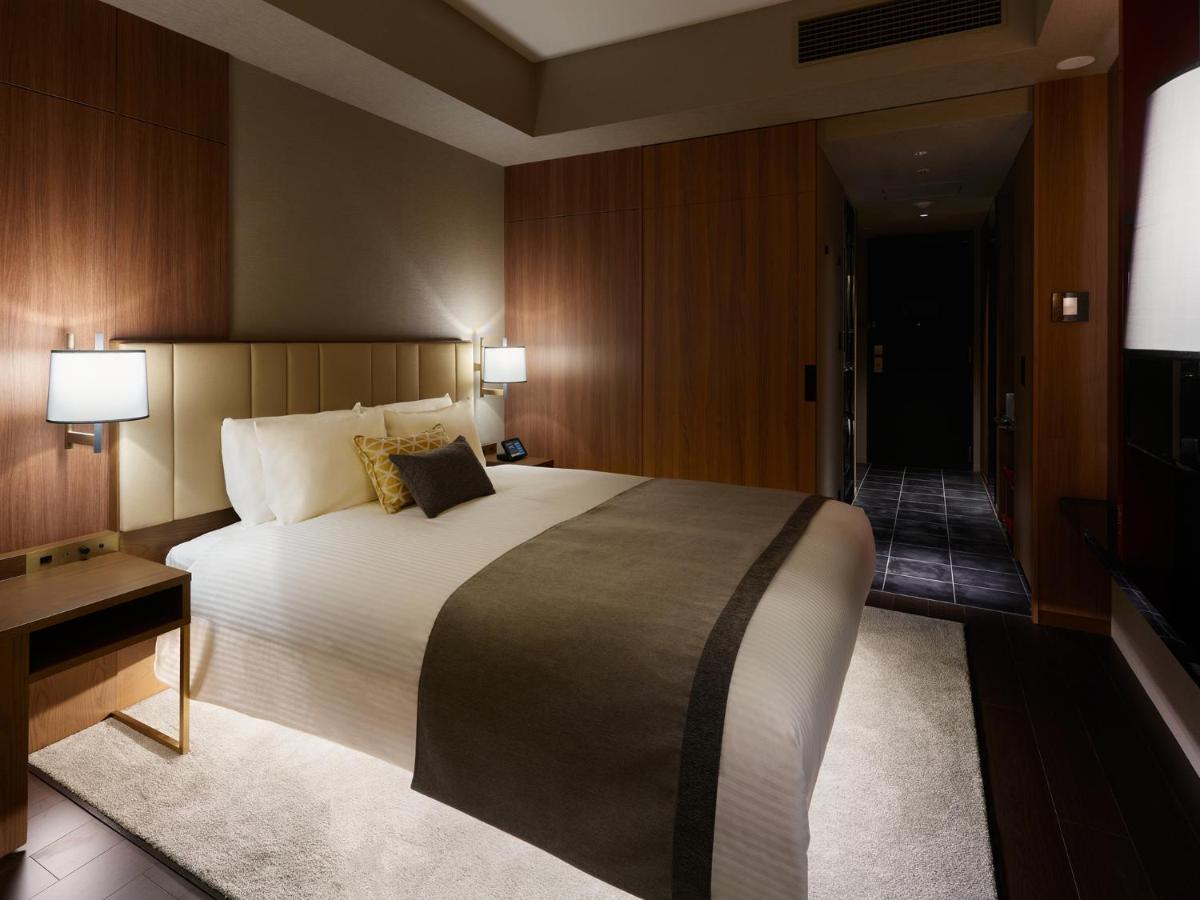 Mitsui Garden Hotel Roppongi Tokyo Premier Esterno foto The image shows a modern hotel room with a warm and elegant design. In the center, there is a neatly made bed with white linens and a dark gray throw blanket. Flanking the bed are two bedside lamps providing soft lighting. The walls have a rich wood 