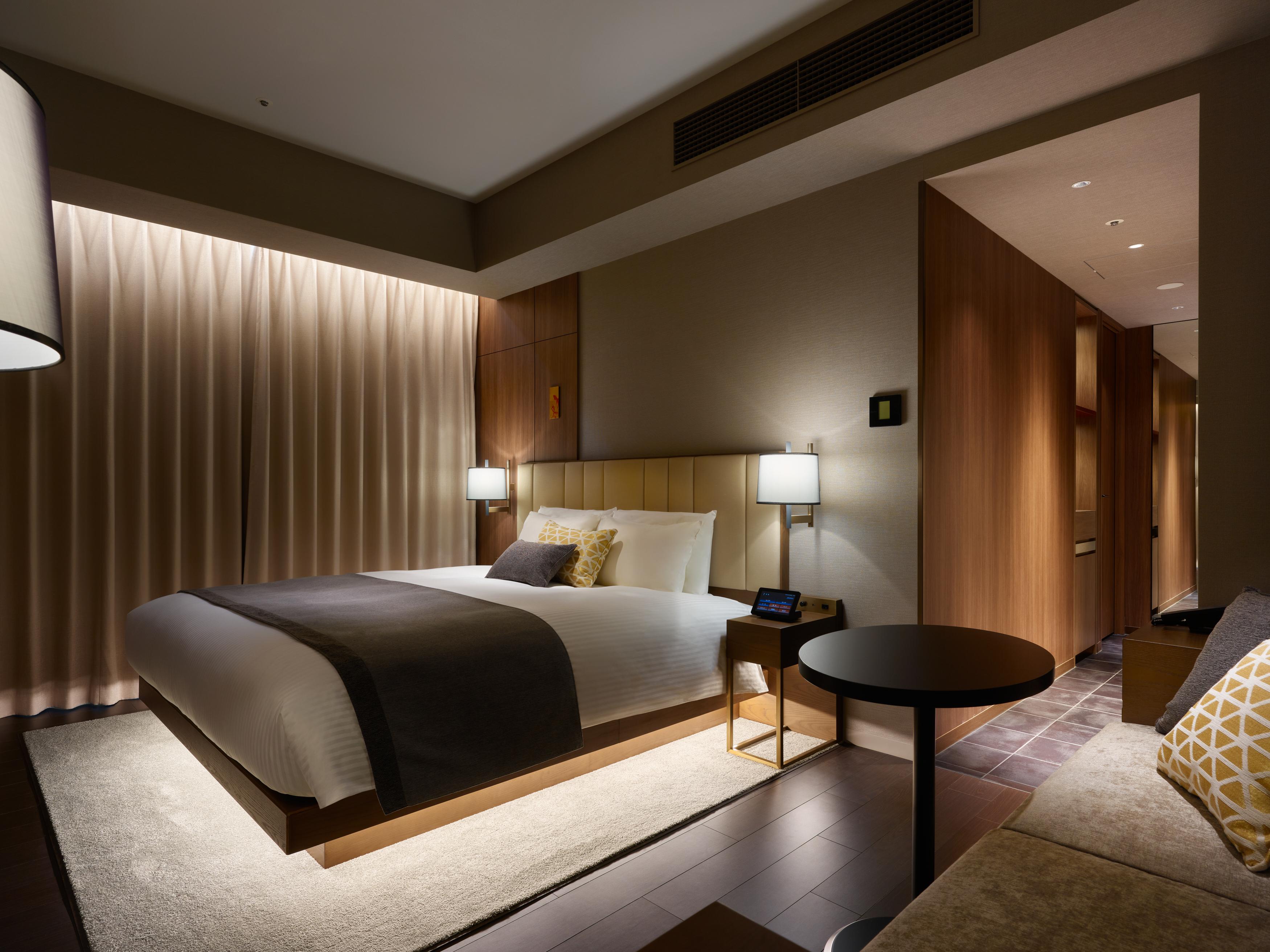 Mitsui Garden Hotel Roppongi Tokyo Premier Esterno foto The photo shows a modern hotel room featuring a king-sized bed with neutral-colored bedding and accent pillows. There are two bedside lamps on either side of the bed, providing a warm glow. The walls have a wood finish, and there's a soft area rug on
