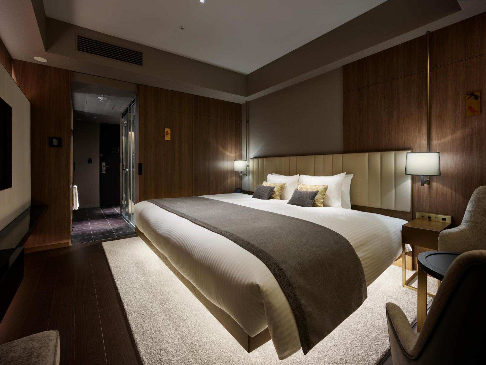 Mitsui Garden Hotel Roppongi Tokyo Premier Esterno foto The photo shows a modern hotel room featuring a large bed with a neutral-colored duvet and a dark throw blanket. The headboard is upholstered, and there are two bedside lamps on either side of the bed. The walls have a warm wood paneling, contributin