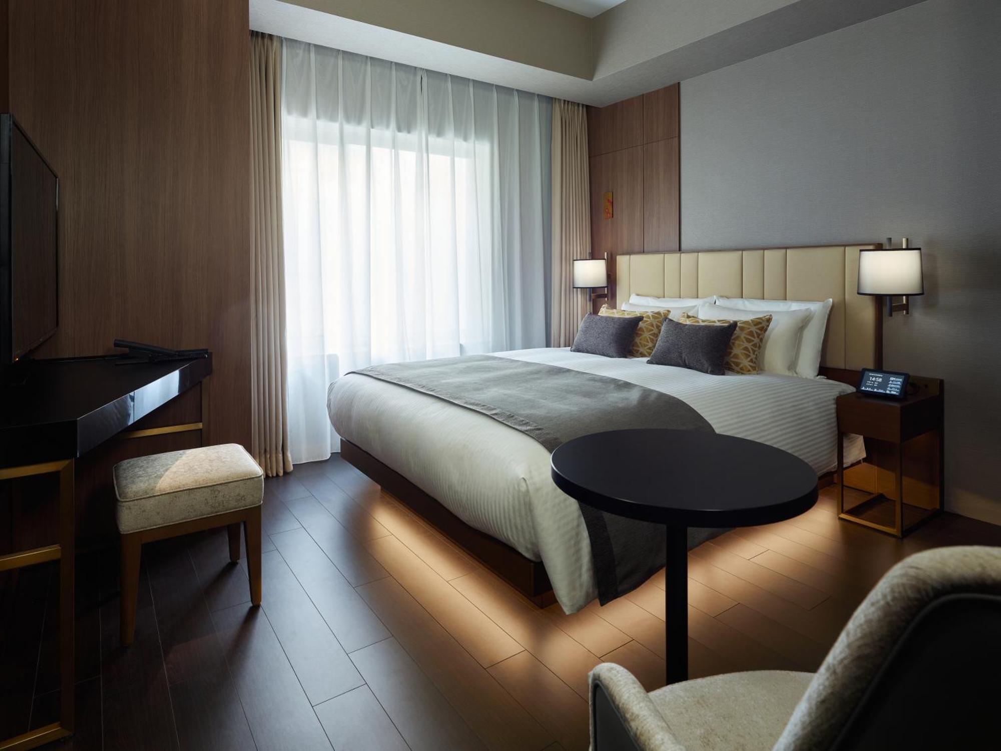 Mitsui Garden Hotel Roppongi Tokyo Premier Esterno foto The photo shows a modern hotel room featuring a large bed with decorative pillows. There are large windows with sheer curtains allowing natural light to filter in. To one side, there is a sleek desk with a chair and a small round table in the center 