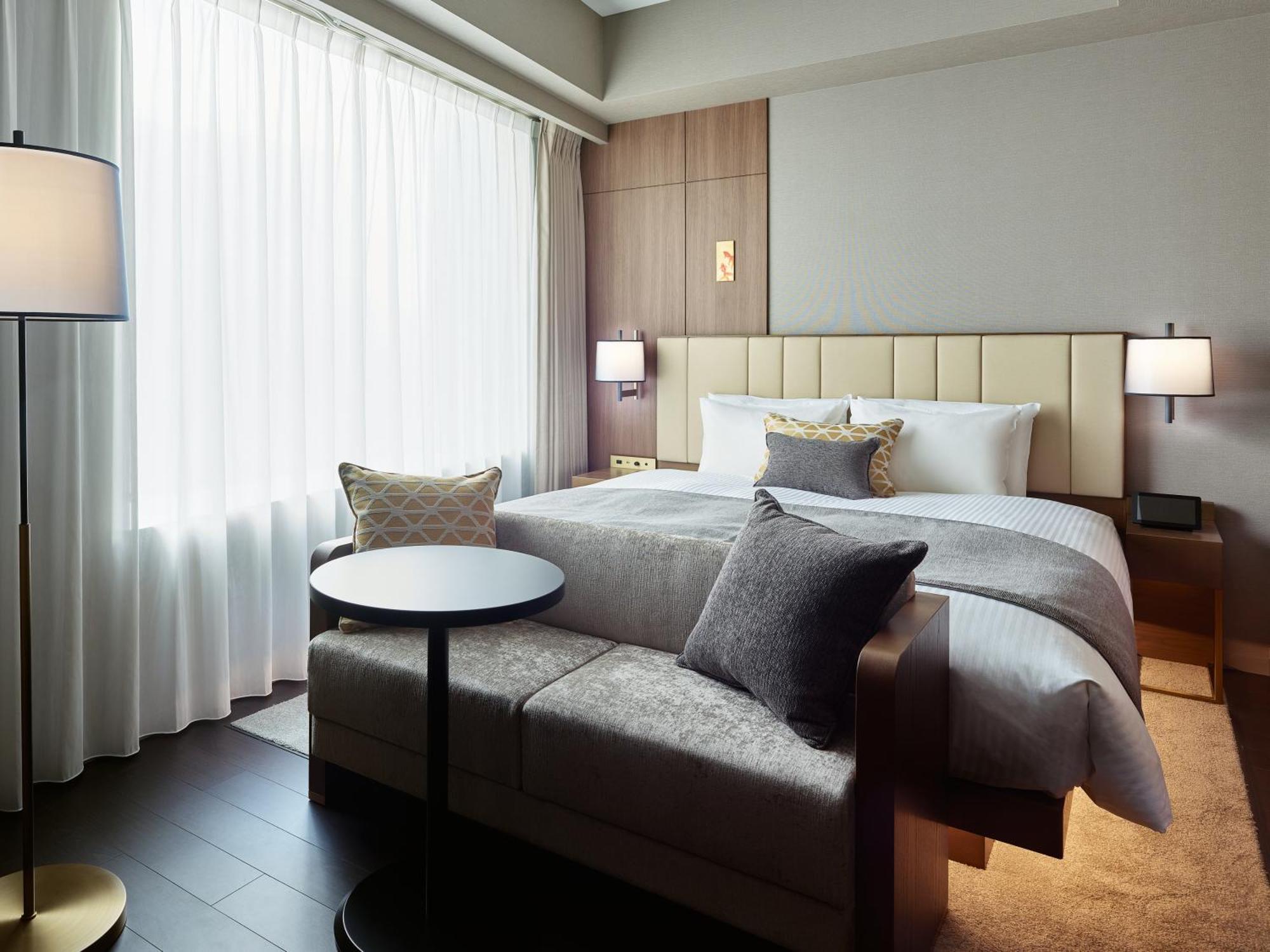 Mitsui Garden Hotel Roppongi Tokyo Premier Esterno foto The photo shows a modern hotel room that features a large bed with a padded headboard and soft bedding. There's a small, round table accompanied by a couch or seating area, adorned with decorative pillows. To the left, a floor lamp provides additiona