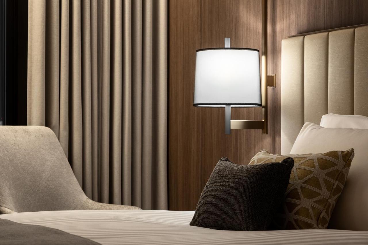 Mitsui Garden Hotel Roppongi Tokyo Premier Esterno foto The photo shows a cozy hotel bedroom setting. There is a neatly made bed with white linens and a few decorative pillows, including a dark one and another with a geometric pattern. To the side, there is a soft armchair and a modern wall lamp mounted a